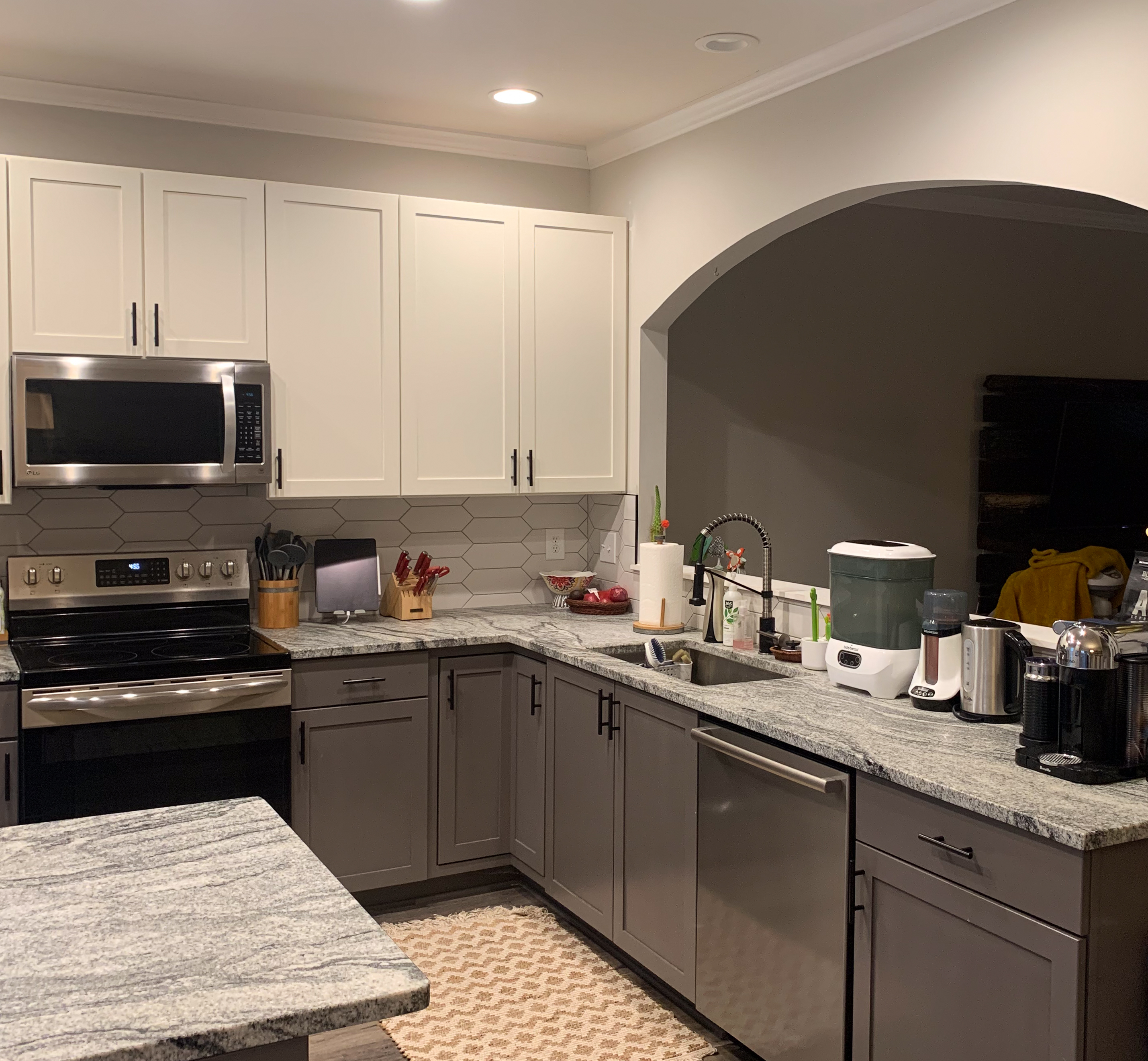 This is a Shaker-style kitchen with an arched opening to the living room. The cabinets are off-white on the top and a deep greige on the bottom.