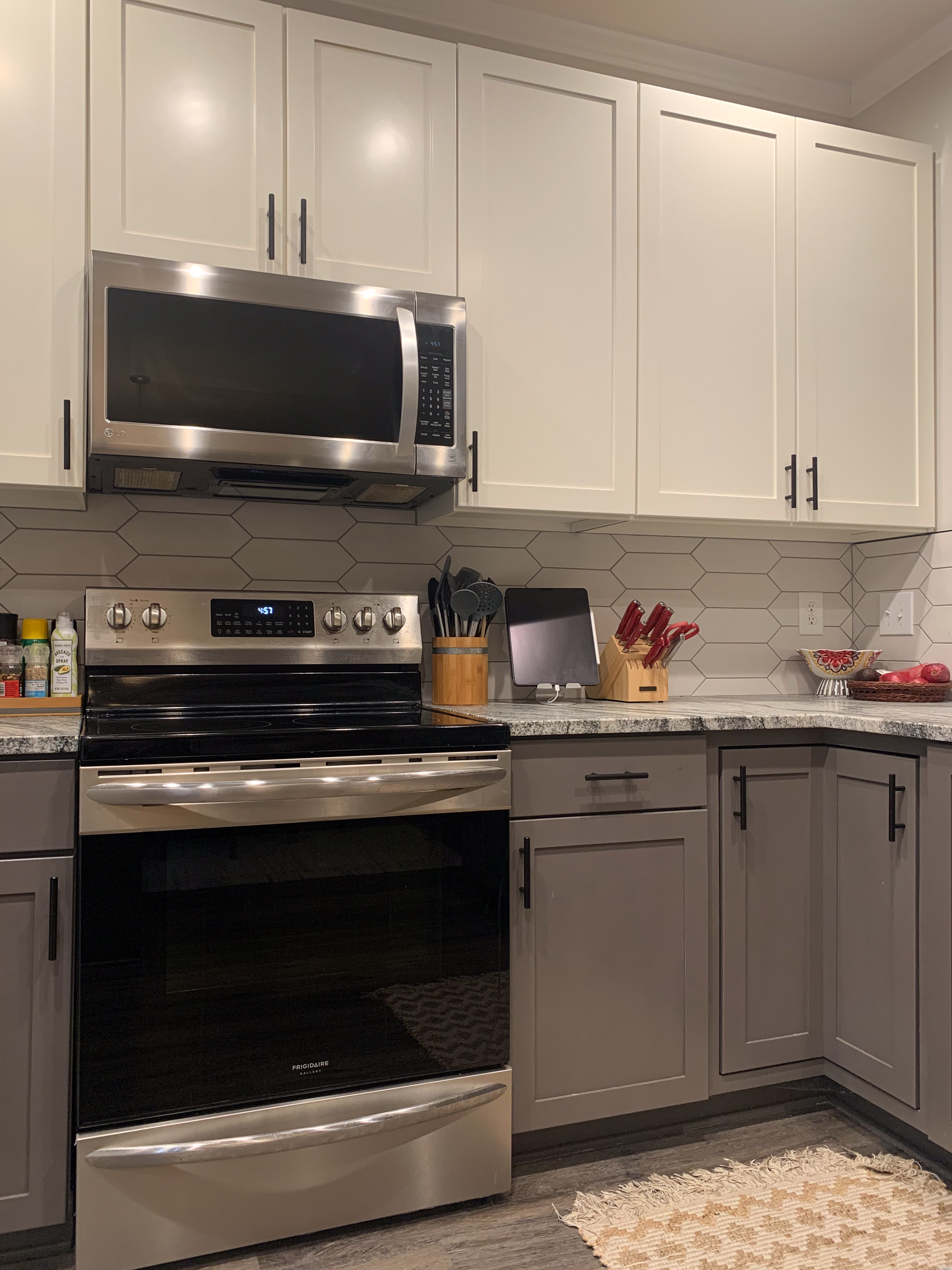 A close up shot of a stainless steel stove and microwave in a Shaker-style kitchen with granite countertops and picket-shaped backsplash tile. The cabinets are off-white on the top and a deep greige on the bottom.