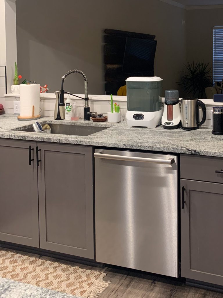 Modern kitchen countertop with granite surface featuring a stainless steel sink and pull-down faucet. Various appliances are displayed, including a white kitchen appliance (possibly a food processor), electric kettle, and blender. Gray cabinets below and a dishwasher are visible. Decorative items like a small cactus plant and paper towel holder add personal touches.