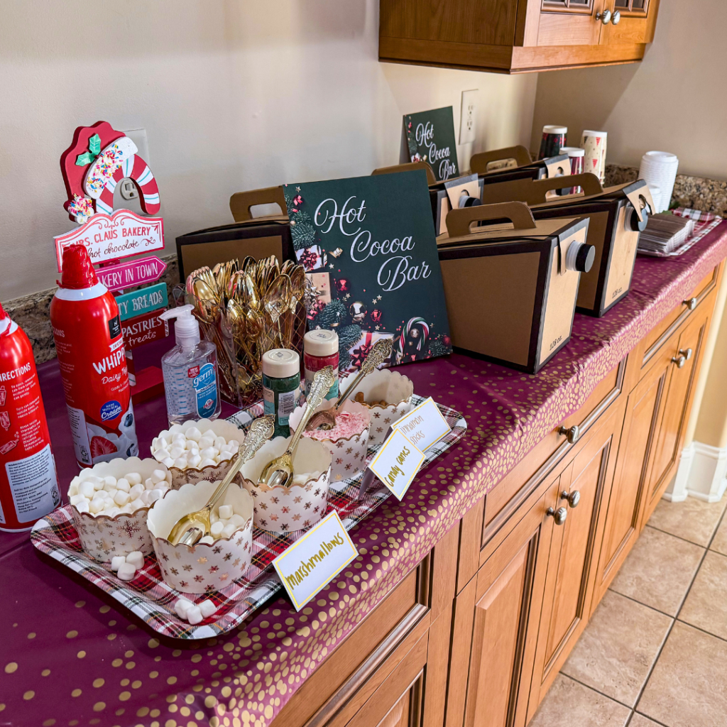 Christmas-themed hot cocoa bar setup featuring decorative signs, bowls of marshmallows, sprinkles, whipped cream, and containers of hot cocoa from Ben & Jerry's.