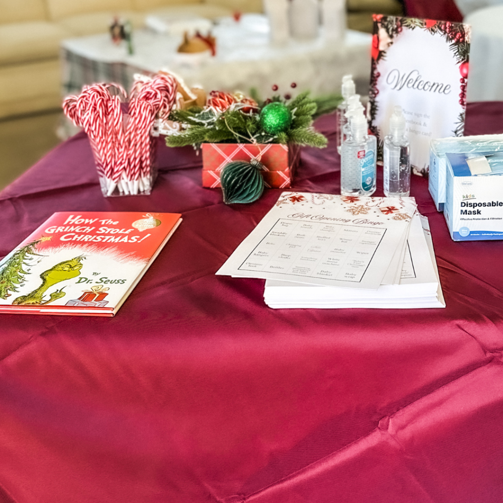 Welcome table at a Christmas baby shower featuring 'How the Grinch Stole Christmas' book, candy cane pens, hand sanitizer, disposable masks, and baby shower games on a burgundy tablecloth.