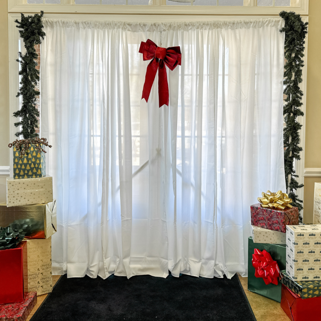 Elegant white curtain photo backdrop framed with garlands, topped with a red velvet bow, and surrounded by beautifully wrapped Christmas gifts in festive patterns.