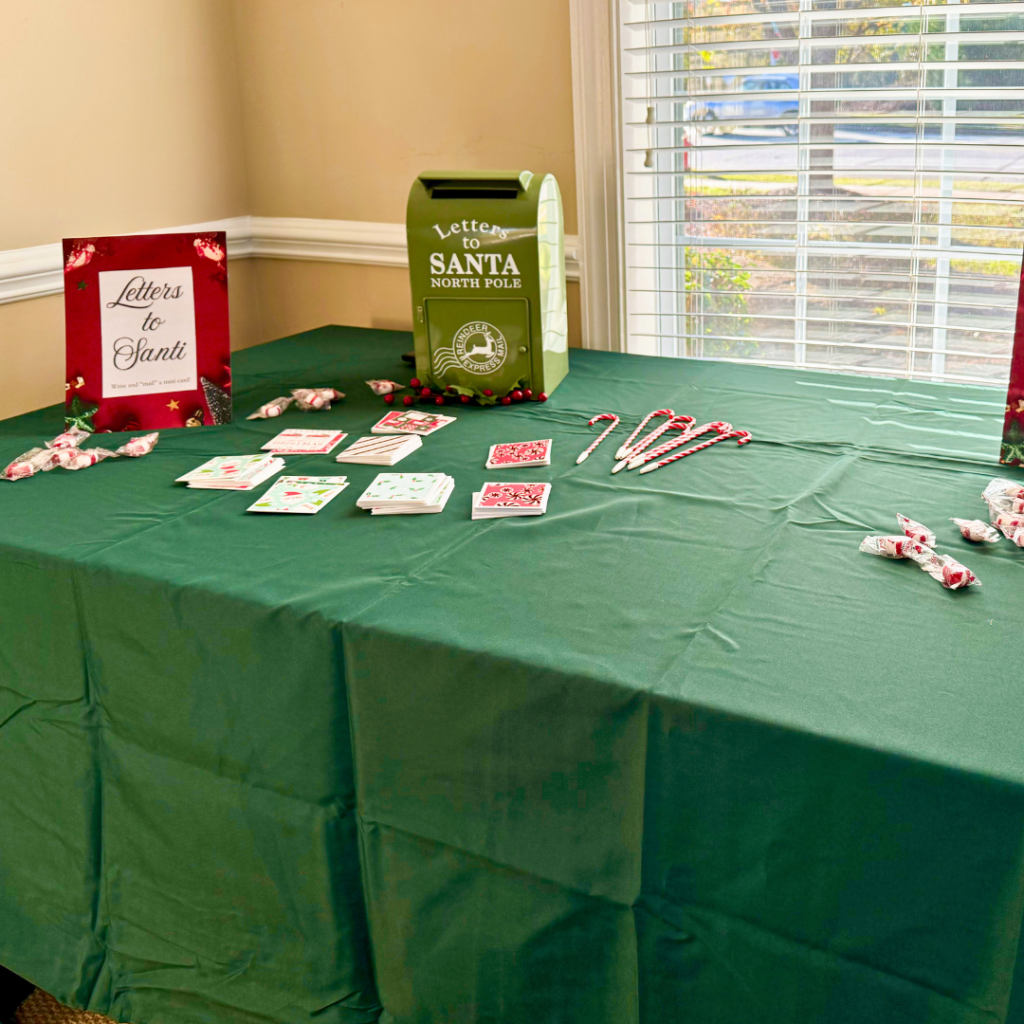 Letters to Santa activity table with a green tablecloth, a green 'Letters to Santa' mailbox, Christmas cards, candy cane pens, and a festive sign encouraging guests to write a note.