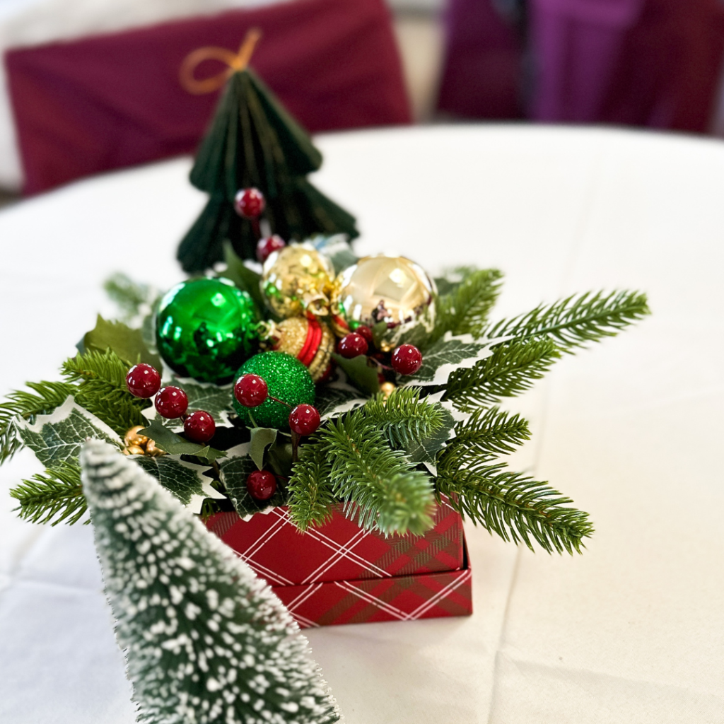 Traditional Christmas centerpiece with evergreen sprigs, red holly berries, gold and green ornaments, and a red plaid gift box base on a white tablecloth.