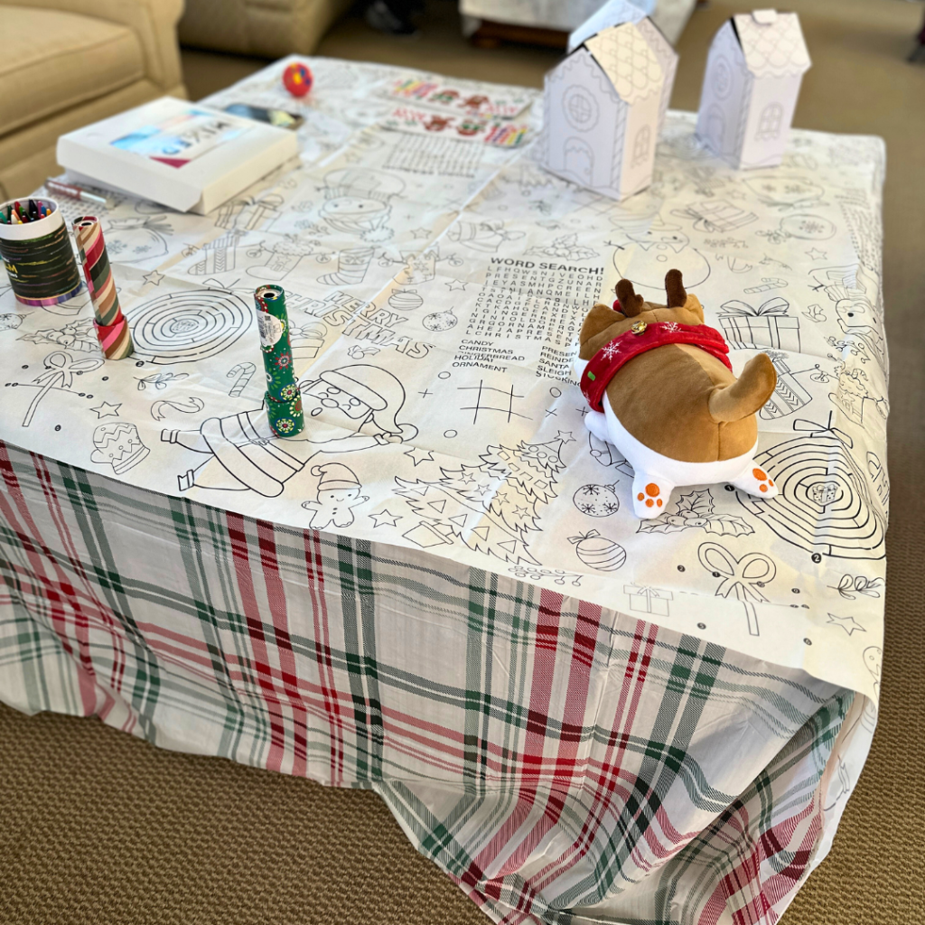 Kids' Christmas activity table featuring a coloring tablecloth with festive designs, kaleidoscopes, crayons, and cardboard gingerbread houses.