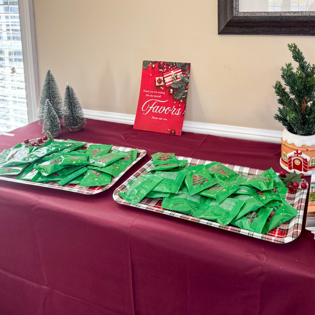 Christmas-themed favor table with green treat bags displayed on plaid-lined trays, miniature Christmas trees, and a festive red sign reading 'Thank you for making this day special. Favors, please take one.