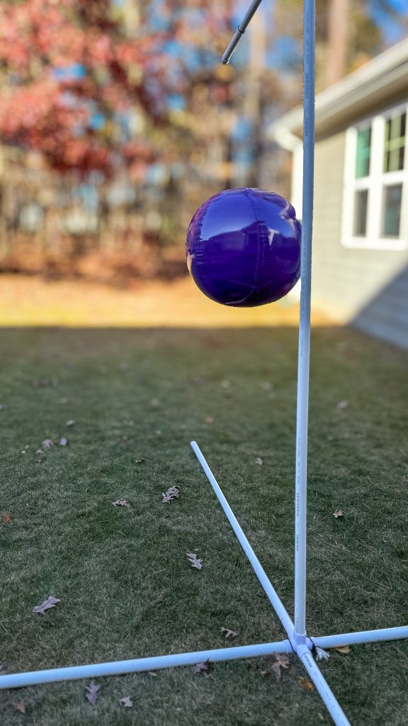A large, glossy purple inflatable ball suspended from a simple white PVC stand in a grassy yard. The ball has been recently spray painted, with autumn leaves scattered on the ground.