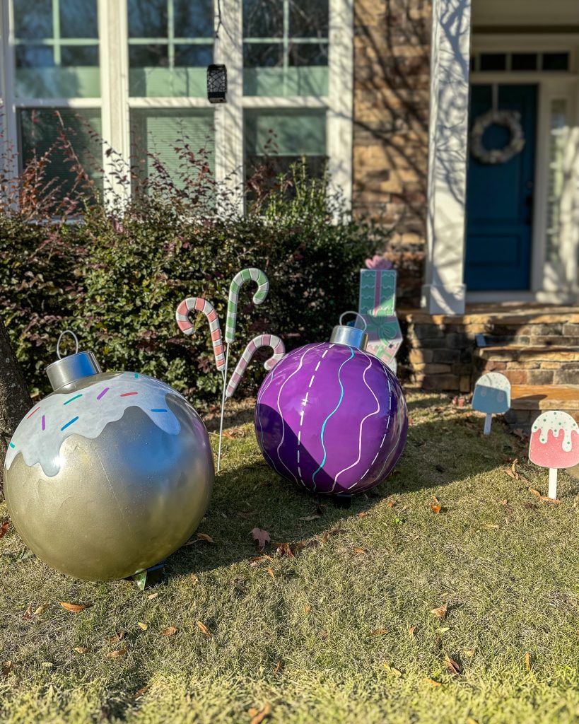 Two oversized DIY outdoor ornaments in a festive yard display. One is metallic silver with white frosting and colorful sprinkles, while the other is glossy purple with neon squiggles. The ornaments are surrounded by pastel candy canes and a wrapped gift decoration.