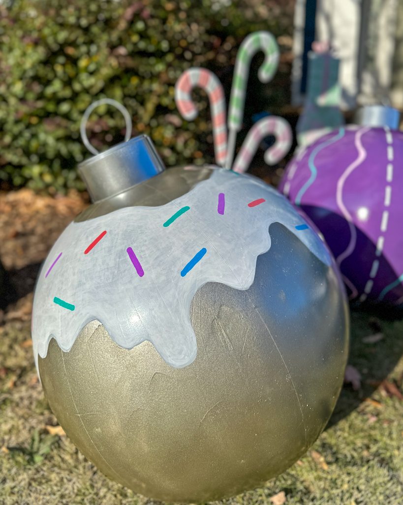 Close-up of a metallic silver oversized ornament with a white frosting drip design and colorful sprinkles. The ornament is part of a festive outdoor display on a grassy lawn.