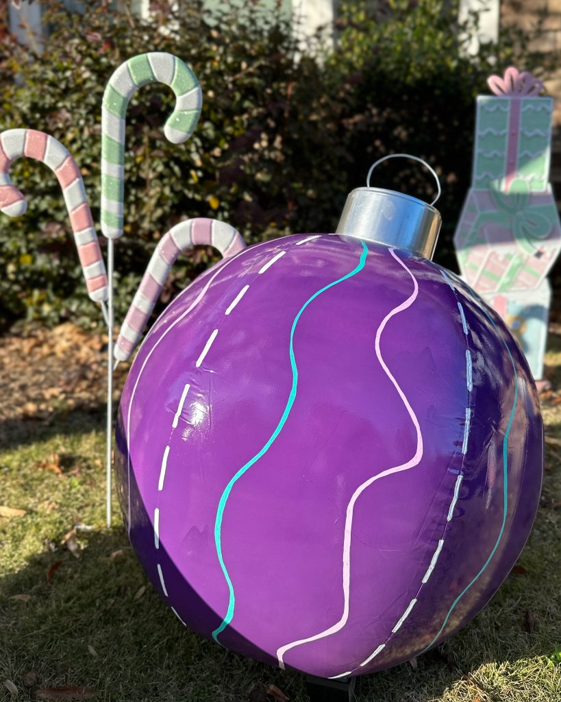 Close-up of a large, glossy purple ornament with pastel squiggles and dashed lines, sitting in a holiday yard display. Pastel candy canes and a wrapped gift decoration are in the background.